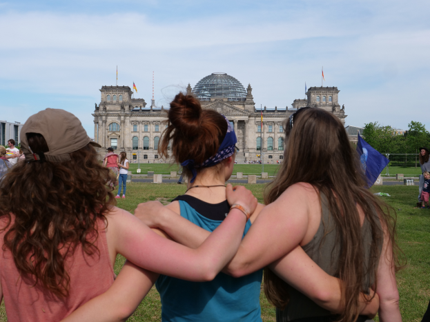 Drei Frauen in verschiedenenfarbigen Kleidern, eine trägt eine Mütze, stehen vor dem Reichstaggebäude in Berlin, Deutschland, mit Menschen, Fahnen, Bäumen, Wolken und Himmel im Hintergrund.