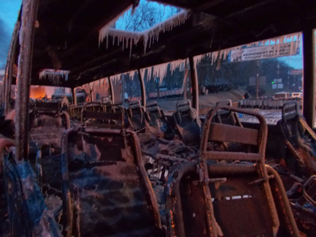 Ein verkohlter, feuerbeschädiger Bus mit Eiszapfen am Dach, vor einem Hintergrund aus Bäumen, Gebäuden und einem klaren Himmel.