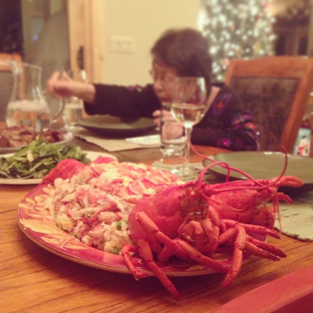Eine Frau sitzt an einem Tisch und hält Chopsticks in der Hand, mit Essen, einem Wasserglas und anderen Gegenständen auf dem Tisch, einem Weihnachtsbaum im oberen rechten Eck und einer Tür mit Steckdosen im Hintergrund.