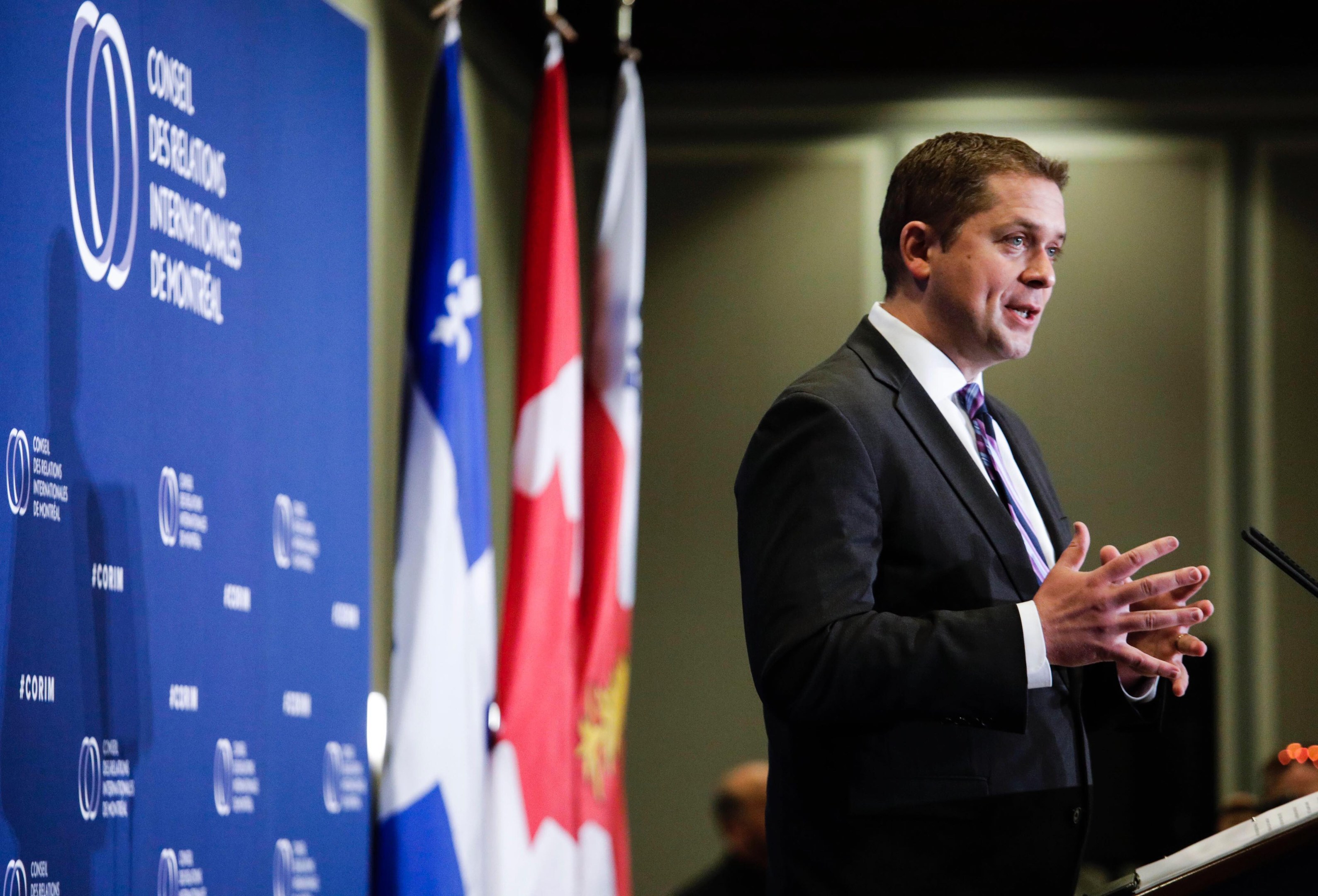 Ein Mann in Anzug und Krawatte steht an einem Rednerpult mit einem Mikrofon vor kanadischen Flaggen und einem Banner, mit ein paar Menschen im Hintergrund.