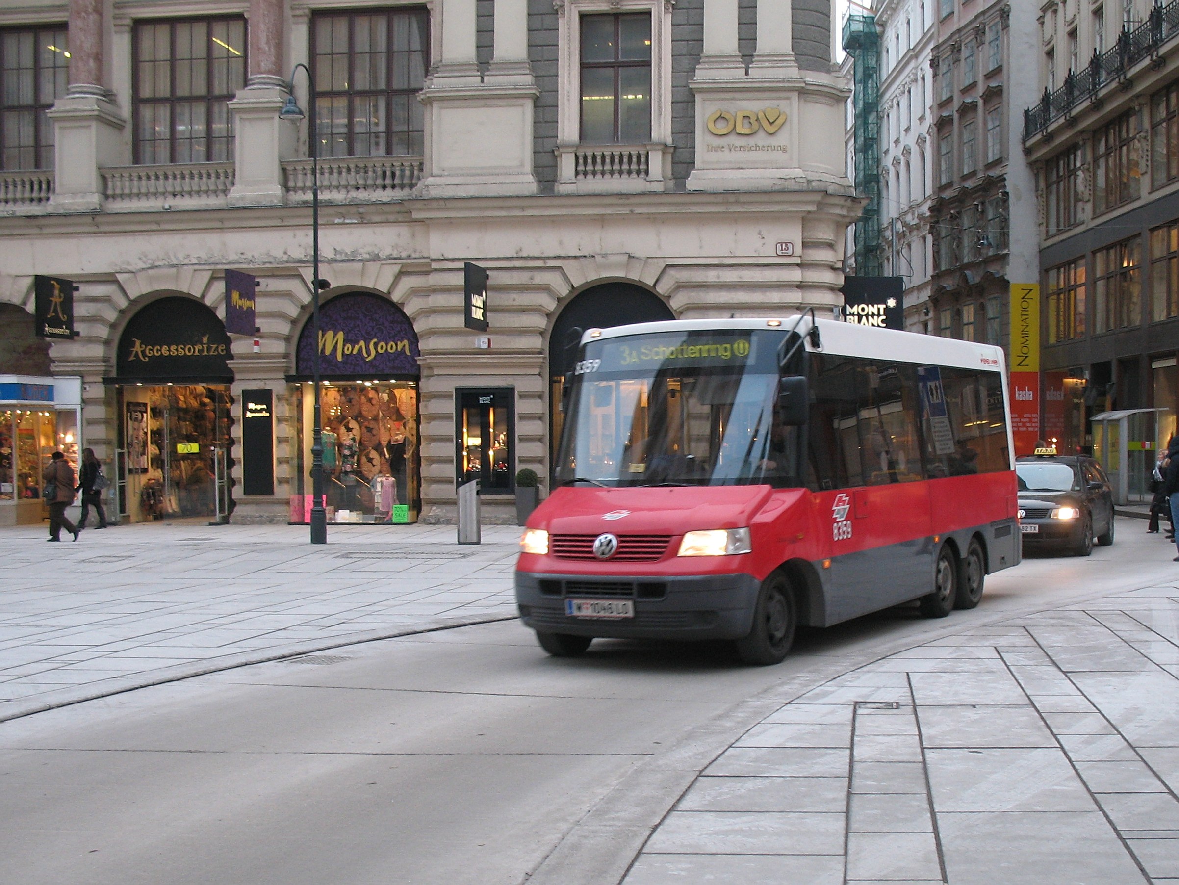 Roter Shuttlebus fährt auf einer von Hochhäusern gesäumten Stadtstraße, Passanten auf dem Gehweg, Laternen, Geschäfte mit Schildern, Himmel im Hintergrund.