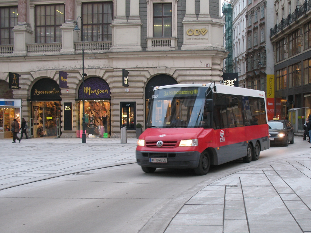 Roter Shuttlebus fährt auf einer von Hochhäusern gesäumten Stadtstraße, Passanten auf dem Gehweg, Laternen, Geschäfte mit Schildern, Himmel im Hintergrund.