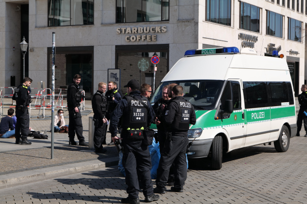 Polizeibeamte vor einem Starbucks-Café stehend mit einem Van auf der rechten Seite und Menschen auf der linken Seite, mit einem Gebäude, einem Schild, einer Straßenlaterne und einem Zaun im Hintergrund.