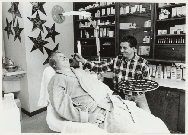 Ein Mann lächelt in einem Zahnarztstuhl, während ein anderer Mann neben ihm steht und ein Tablett hält, mit einem Waschbecken, einer Uhr und Regalen mit Flaschen im Hintergrund.
