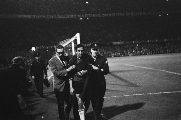Eine Gruppe von Männern, die auf einem Fußballfeld mit einem Torpfosten und beleuchteten Tribünen im Hintergrund feiern, dargestellt in Schwarz-Weiß.
