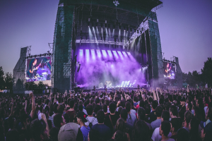 Eine große Menschenmenge auf einem Musikfestival schaut Künstlern auf einer hell erleuchteten Bühne mit Instrumenten und Bildschirmen zu, die vor einem Gebäude, Bäumen und einem klaren blauen Himmel aufgebaut ist.