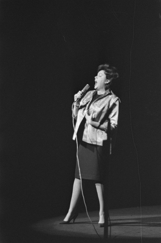 Eine Frau in einem schwarz-weißen Outfit aus den 1950er-Jahren singt fröhlich auf einer von einem Scheinwerfer beleuchteten Bühne in ein Mikrofon, ihr Haar ist im klassischen 1950er-Jahre-Stil frisiert.