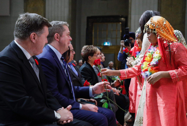 Eine Gruppe von Menschen, die in einem Raum zusammensitzen, mit einer Frau in der Mitte, die einen Kranz trägt und Blumen hält, flankiert von einem Mann im Anzug und jemandem mit einer Kamera, vor einer Wand mit einer Tür.