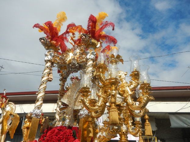 Ein prächtiger goldener und roter Festwagen, geschmückt mit Blumen und anderen Dekorationen, bei einem Karnevalsumzug, mit einem Gebäude, Strommasten mit Kabeln und einem bewölkten Himmel im Hintergrund.