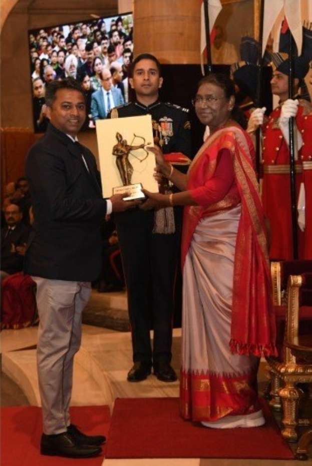 Ein Mann in einem schwarzen Blazer, weißem Hemd und grauen Hosen hält eine Trophäe, während er neben einer Frau in einem aschgrauen Sari auf einem roten Teppich an einer Preisverleihung steht, mit Menschen, Stühlen, Flaggen und einem Bildschirm im Hintergrund.