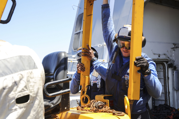 Zwei Männer in blauen Uniformen und Schutzausrüstung arbeiten an einem gelben Gabelstapler, einer hält ein Metallobjekt, neben einem Fahrzeug unter einem Himmel.