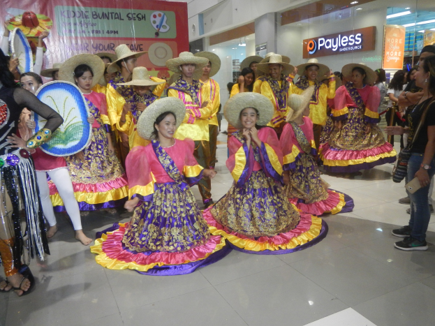 Eine Gruppe von Menschen in bunten Kleidern und Hüten, die auf einem Einkaufszentrum während eines festlichen Ereignisses tanzen, während andere Musikinstrumente halten und ein Banner im Hintergrund sichtbar ist.