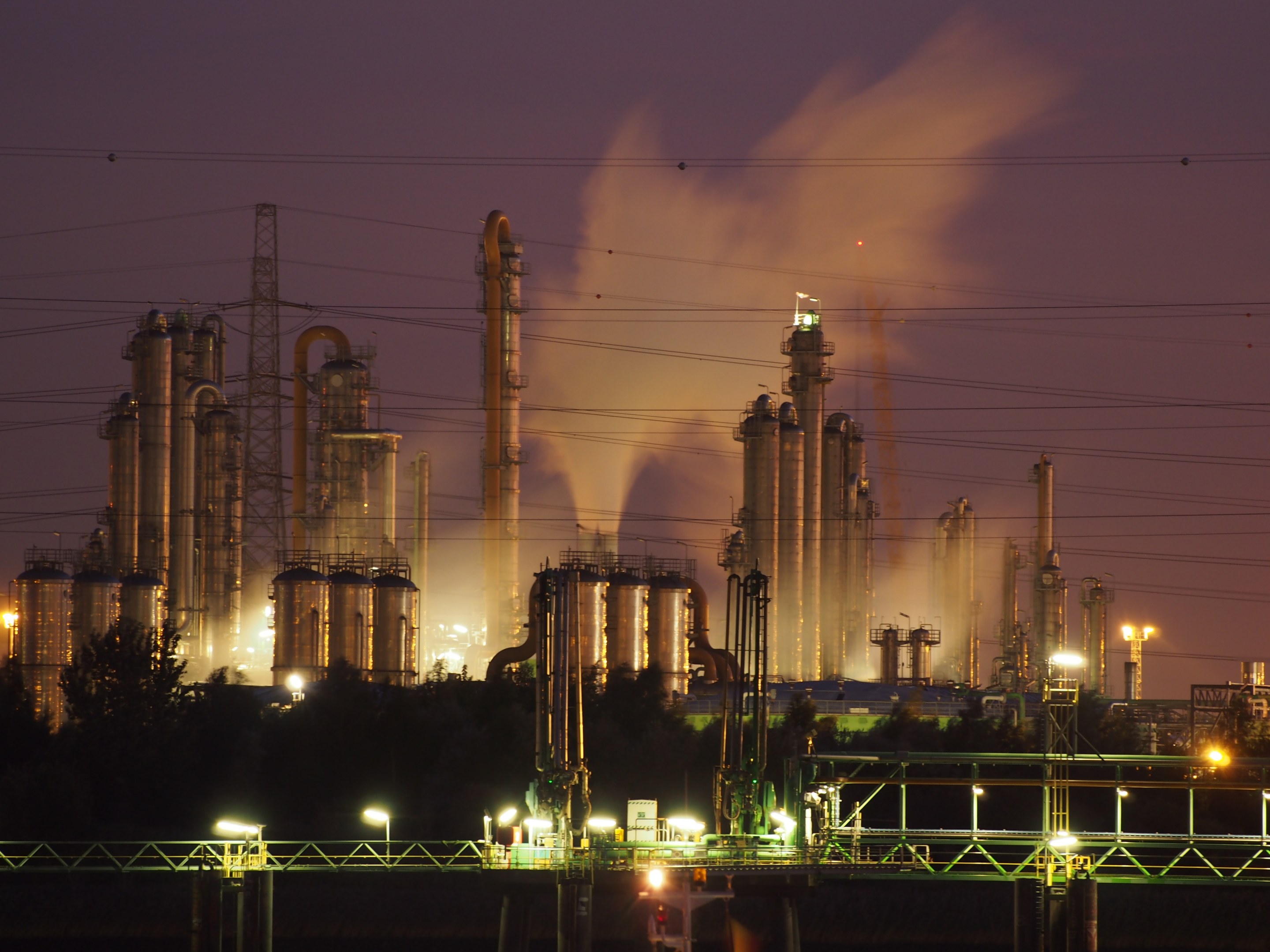 Ölraffinerie bei Nacht mit Rauchwolken, beleuchtet von umliegenden Lichtern, vor einem sichtbaren Himmel, mit einem Wasserzeichen.