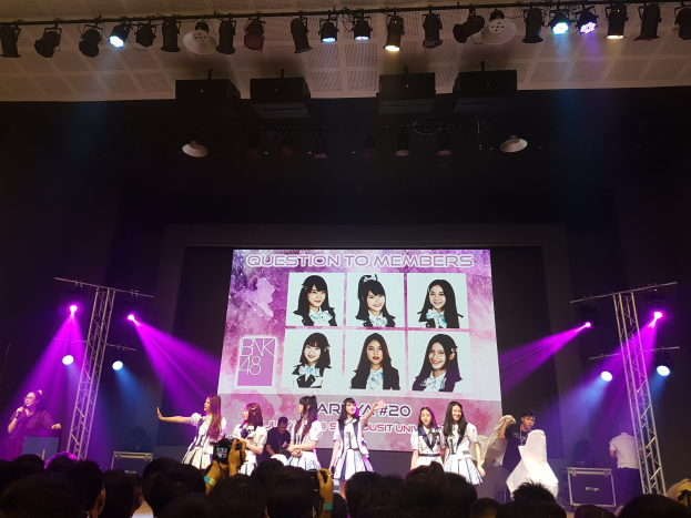 Eine Gruppe von Menschen performs auf einer hell beleuchteten Bühne mit Trägern, während eine Menge darunter das Ereignis aufzeichnet; ein großer Bildschirm im Hintergrund zeigt Bilder der K-Pop-Gruppe BK48.