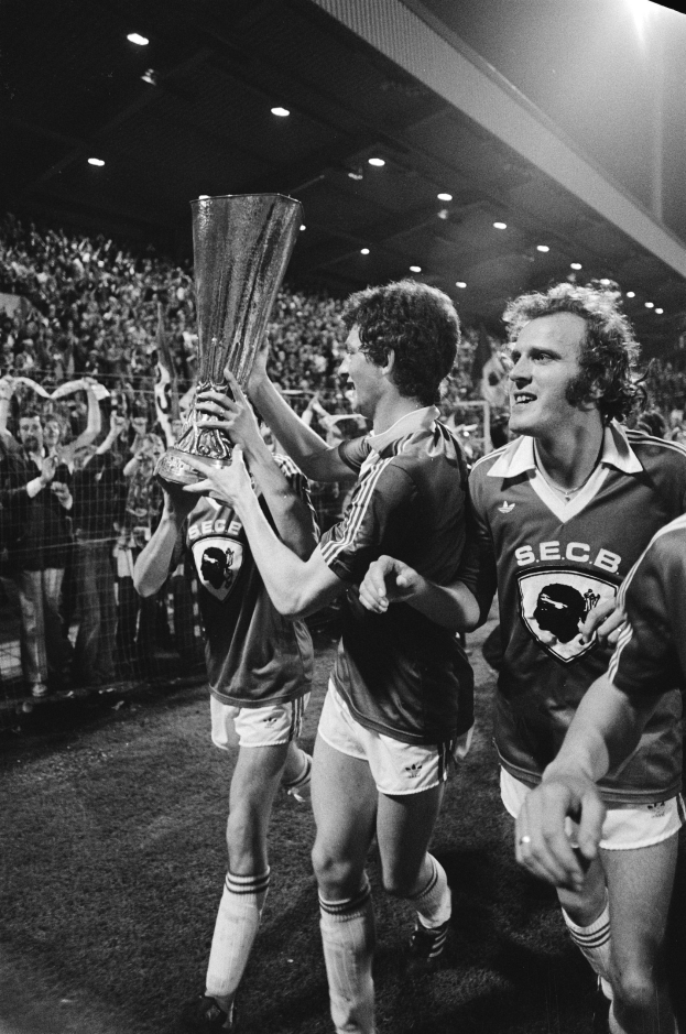 Schwarzes und weißes Foto von Fußballspielern, die einen Pokal auf einem Feld halten und ihren Sieg im FA-Cup-Finale feiern, während Zuschauer hinter einem Zaun stehen und von Stadionlichtern beleuchtet werden.