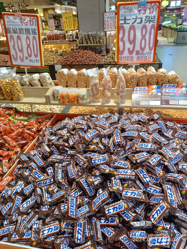 Eine Ladenvitrine mit verschiedenen Süßigkeiten, darunter Snickers-Riegel, und anderen Lebensmitteln auf Regalen im Hintergrund.