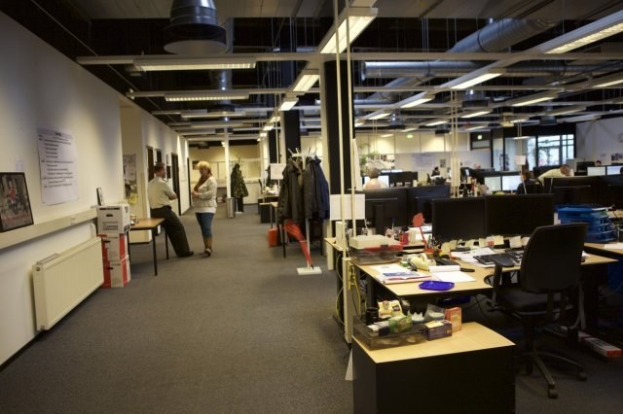 Großes Großraumbüro mit Menschen an Schreibtischen, Computern und Monitoren, umgeben von Stühlen, Papieren, Büchern und hängenden Kleidungsstücken, mit Wandtafeln und Deckenlampen.