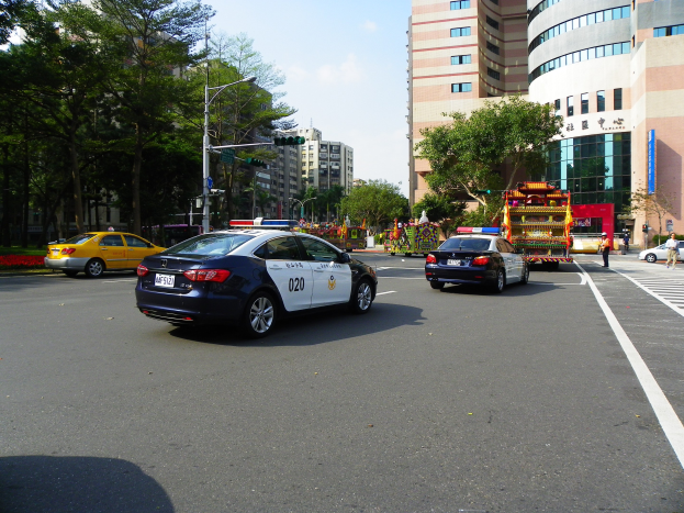 Polizeiauto fährt auf einer Straße mit hohen Gebäuden, Bäumen, Laternen, Ampeln und Menschen unter einem bewölkten Himmel.