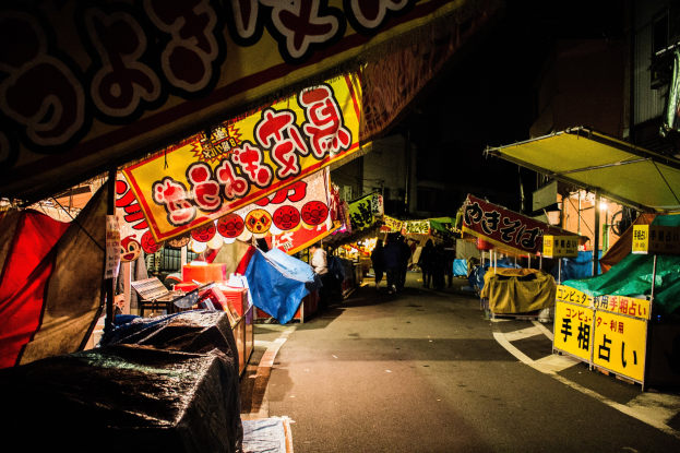 Ein belebter Nachtmarkt mit beleuchteten Ständen, die eine Straße säumen, Menschen, die zwischen ihnen hindurchgehen, und eine zentrale Figur, die von der Aktivität umgeben ist.