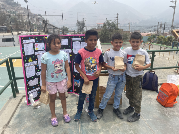 Gruppe von Kindern auf einem Basketballfeld, die Papiertütten halten, mit Gegenständen auf der rechten Seite und einer Tafel mit Text auf der linken Seite, vor dem Hintergrund von Zäunen, Masten, Straßlaternen, Bäumen, Gebäuden, Höhen und Himmel.