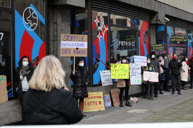 Gruppe von maskierten Menschen mit Schildern und Plakaten vor einem Glasfrontgebäude, mit Kameras und einem Hund, wahrscheinlich protestierend gegen ein deutsches Maskenverbot in Schulen.