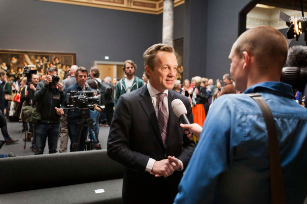 Ein Mann im Anzug und Krawatte wird von einem Reporter interviewt, während er vor einer Menge steht, einige halten Kameras, mit einem Sofa im Vordergrund und einer Wand mit einem Foto Rahmen, Lampen und einer Säule im Hintergrund.