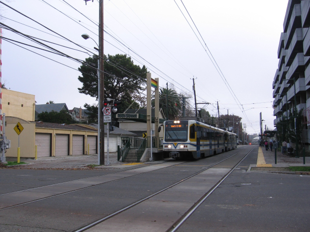 Straßenbahn fährt durch eine Stadtstraße mit hohen Gebäuden, Strommasten, Fußgängern, Bäumen und einem klaren Himmel.