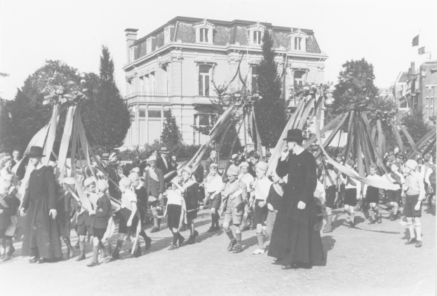 Schwarzes und weißes Bild einer Gruppe von Menschen, die vor einem Gebäude auf einer Straße gehen, einige halten Fahnen, mit Hüten, Bäumen, Gebäuden und einem Maibaum im Hintergrund.
