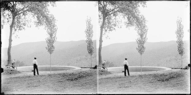 Schwarz-weißes Foto eines Mannes, der vor einem Teich steht, umgeben von Bäumen, Hügeln und einem klaren Himmel, mit üppiger Vegetation um den Teich herum.