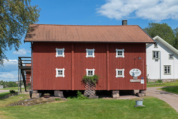 Ein rotes Haus im Vordergrund mit einem weißen Haus dahinter, das auf grünem Gras mit Bäumen steht, unter einem bewölkten Himmel, mit einer Tafel mit Text.