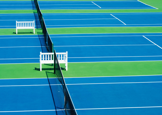 Blauer und grüner Tennisplatz mit zwei weißen Bänken, einem Netz in der Mitte und grünem Rasen drumherum.