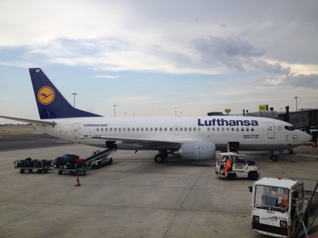 Ein Lufthansa Airbus A320-200 ist auf dem Frankfurter Flughafen geparkt, mit verschiedenen Bodenfahrzeugen, Wagen, Verkehrskegeln, Pfosten und Texttafeln im Hintergrund unter einem bewölkten Himmel.
