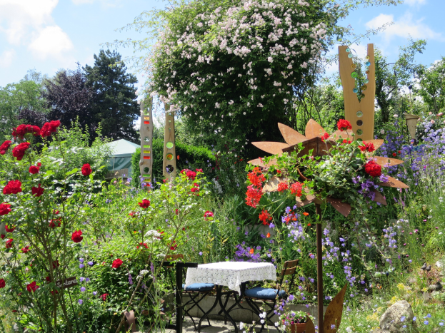Ein Garten mit bunten Blumen und Pflanzen, der einen Tisch mit einer Decke und Stühlen in der Mitte und umgeben von Bäumen unter einem bewölkten Himmel bei der Chelsea Flower Show zeigt.