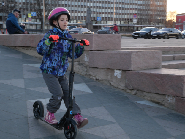 Ein junger Junge mit Helm und Handschuhen fährt auf einem Gehweg einen Roller, mit Treppen, Fahrzeugen, Menschen, Bäumen, Pfählen, Brettern, Gebäuden und einem klaren blauen Himmel im Hintergrund.