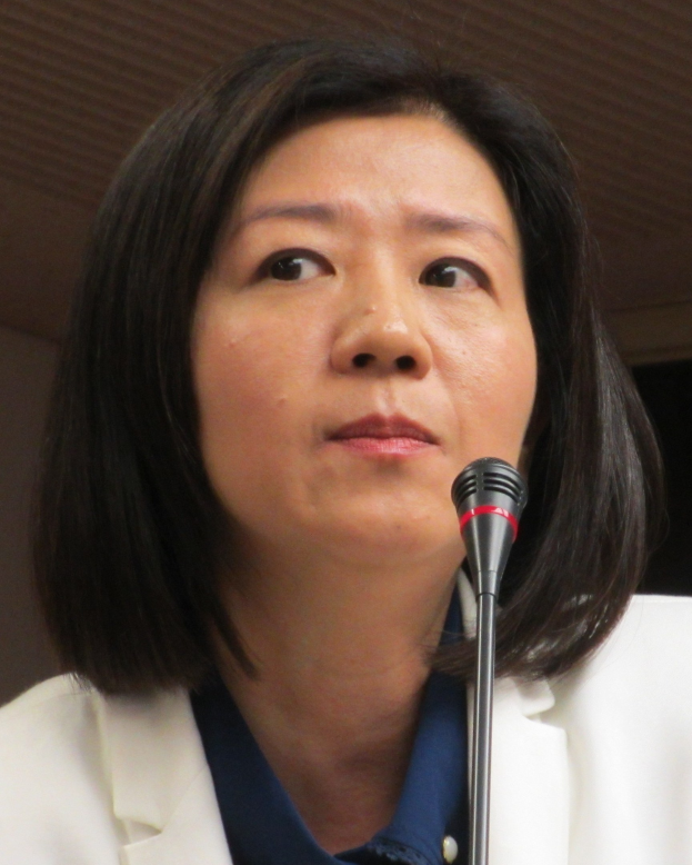 Frau in weißer Jacke, die in ein Mikrofon spricht, trägt einen Anzug mit ernster Miene vor einer Wand.