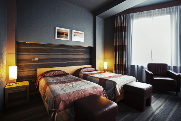 Hotelzimmer mit zwei Betten, jeweils mit Decken und Kissen, zwei Hockern, einem Stuhl, zwei Tischen mit Lampen, zwei gerahmten Fotos an der Wand und einem Fenster mit Vorhängen.