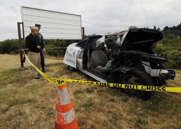 Ein Polizeibeamter in Uniform steht neben einem schwer beschädigten Auto am Straßenrand mit eingedellter Motorhaube und zersplitterten Fenstern unter einem bewölkten Himmel.
