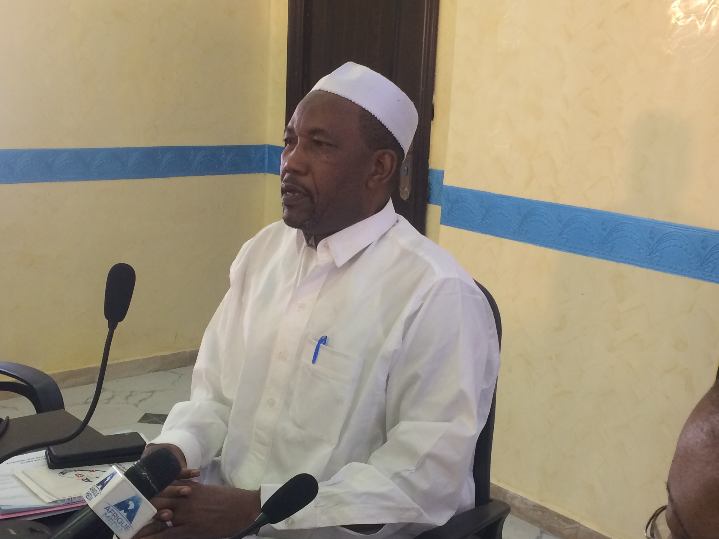 Ein Mann in weißem Anzug und Hut sitzt an einem Tisch mit Mikrofonen, spricht, während Papiere und Gegenstände auf dem Tisch zu sehen sind, mit einer anderen Person mit Brille zu seiner Rechten, in einem Raum mit einer Wand und einer Tür im Hintergrund.