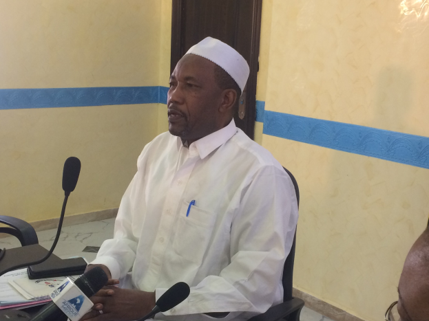 Ein Mann in weißem Anzug und Hut sitzt an einem Tisch mit Mikrofonen, spricht, während Papiere und Gegenstände auf dem Tisch zu sehen sind, mit einer anderen Person mit Brille zu seiner Rechten, in einem Raum mit einer Wand und einer Tür im Hintergrund.