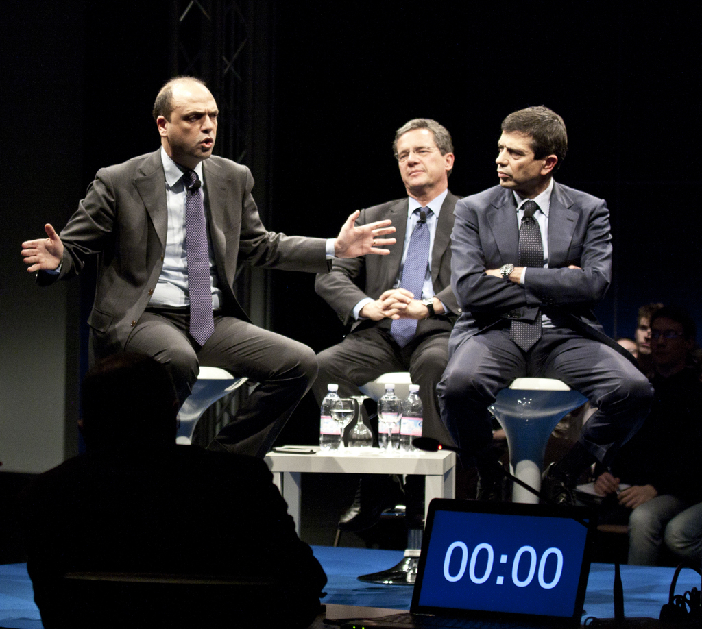 Drei Männer in Anzügen sitzen auf Stühlen, wobei der Mann links spricht und die anderen beiden zuhören; ein kleiner Tisch mit drei Flaschen davor, ein Laptop unten im Bild und ein paar weitere Menschen rechts, die die Männer anschauen.