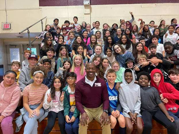 Gruppe von Schülern, die für ein Foto in einer Turnhalle posieren, wobei einige auf der Treppe sitzen und andere stehen, eine Tür und ein Ausgangsschild links sichtbar