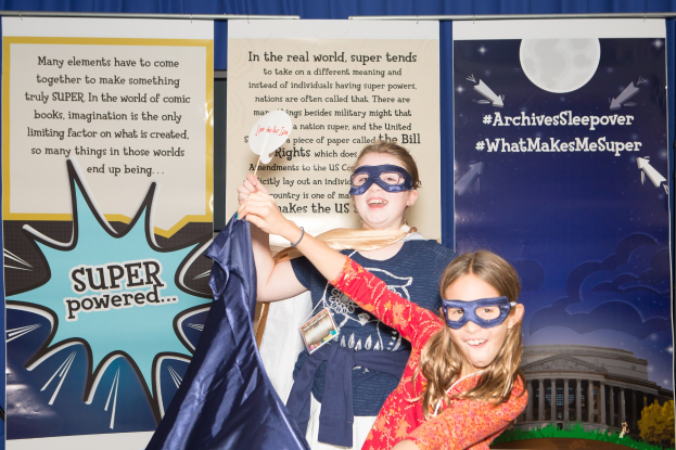 Zwei junge Mädchen in Superheldenkostümen posieren für ein Foto vor einem blauen Vorhang bei einem Schulereignis, eine hält ein Tuch und beide tragen Masken.