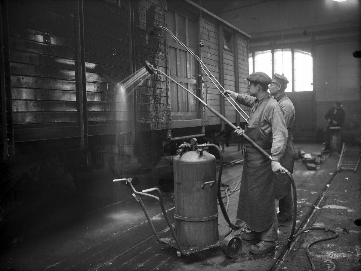 Zwei Männer in einer Fabrik sprühen Wasser auf einen Zug, während sie Rohre halten und ein Zylinder und ein Wagen in der Nähe stehen.