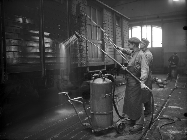 Zwei Männer in einer Fabrik sprühen Wasser auf einen Zug, während sie Rohre halten und ein Zylinder und ein Wagen in der Nähe stehen.
