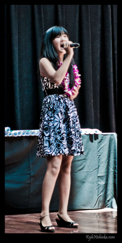 Eine Frau in einem farbenfrohen Kleid singt auf der B├╝hne in ein Mikrofon, mit einem tisch voller Papiere und einem Vorhang im Hintergrund.