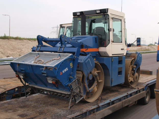 Ein Walzenstapler wird auf einen Tieflader geladen an der Seite der Straße mit Laternenmasten, Bäumen und einem klaren blauen Himmel im Hintergrund.