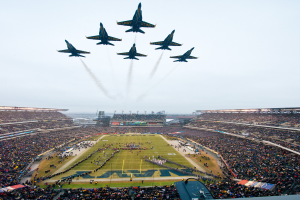 Eine Gruppe von Jets, die in Formation über ein Footballfeld fliegen, mit einem Stadion voller Menschen im Hintergrund und dem Himmel oben sichtbar.