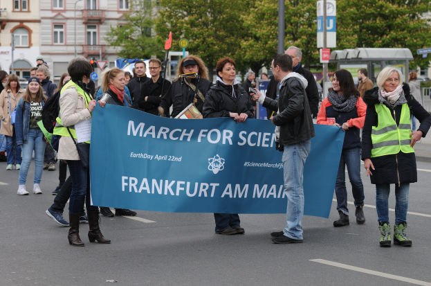 Gruppe von Menschen marschiert die Straße entlang und hält ein "March for Science Frankfurt am Main"-Schild mit Bäumen, Schildern und Gebäuden im Hintergrund unter einem klaren blauen Himmel.