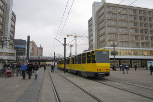 Gelber Straßenbahnwagen auf einer Stadtstraße mit Fußgängern, parkenden Fahrrädern auf dem Gehweg, Gebäuden, Versorgungsmasten, Oberleitungen, einem Baukran und Himmel im Hintergrund.