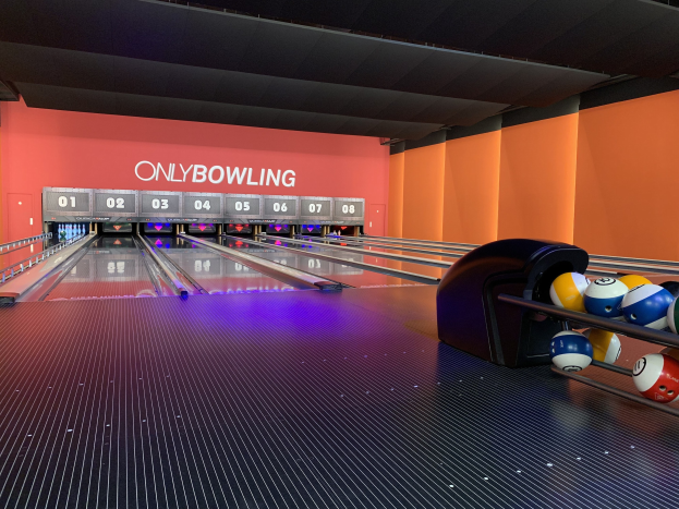 Eine Bowlingbahn mit einer Reihe von Bowlingkugeln auf der rechten Seite, aufgestellte Bowlingkegel in der Mitte und eine Wand mit Text im Hintergrund unter einer sichtbaren Decke.